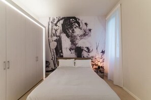 Ein Schlafzimmer mit einem großen Bett, weißen Wänden, einem schwarz-weißen abstrakten Wandbild und einem Nachttisch mit Lampe.