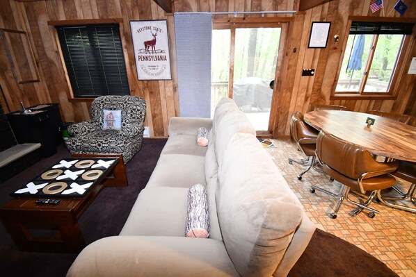 A cozy cabin interior with a sofa, patterned armchair, wooden dining table, and a game of tic-tac-toe.