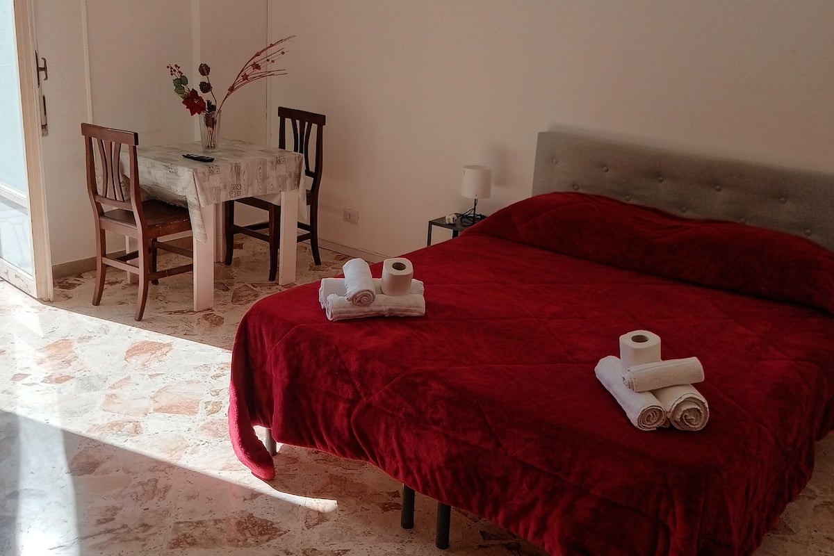 A hotel room with a bed, a table with a vase, and a chair.