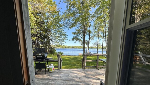 Una terraza de madera con una parrilla y una mesa de picnic, con vista a un lago rodeado de árboles y un muelle.
