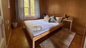 Un dormitorio con paneles de madera, una cama, una ventana con cortinas, una mesita de noche pequeña y una planta en maceta.