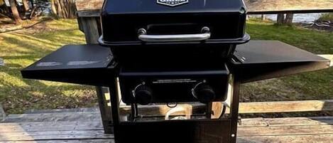 A portable grill on a wooden deck with a table and benches in the background.