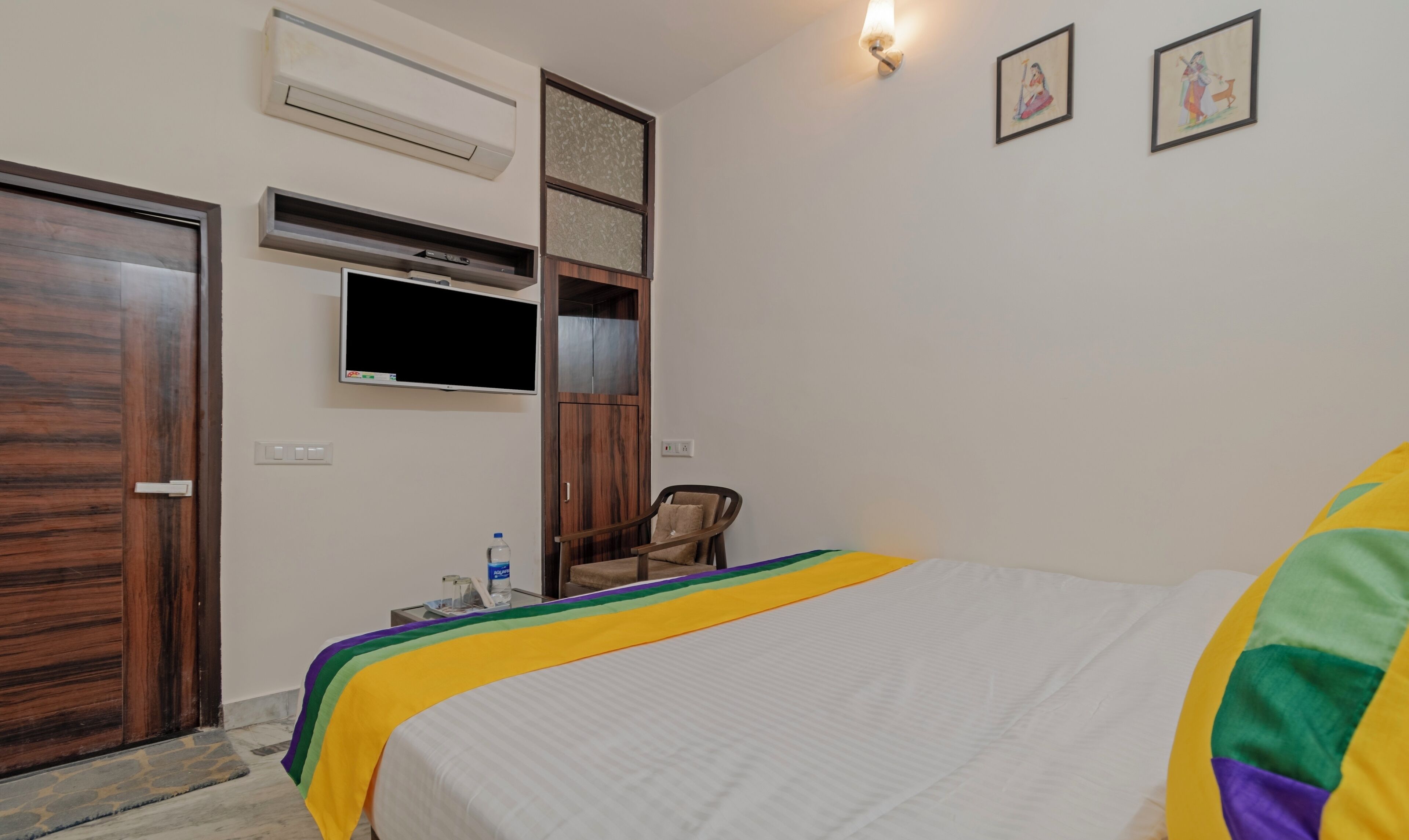 A hotel room with a bed, a TV, an air conditioning unit, a chair, and a water bottle on a small table.