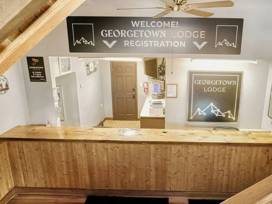 Una recepción con un cartel que dice '¡Bienvenidos! Registro del Georgetown Lodge' y un aviso de vigilancia las 24 horas.