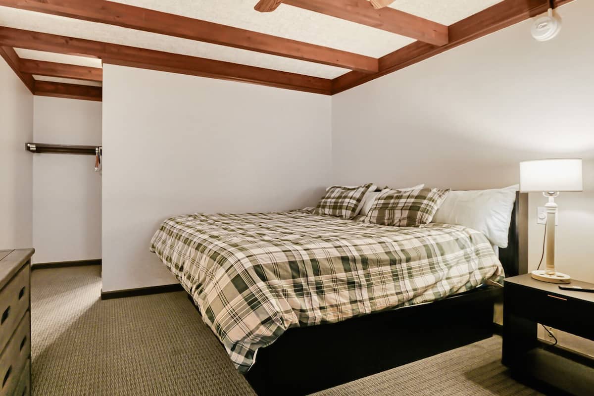 A bedroom with a bed, bedside table, lamp, wooden dresser, and a ceiling with exposed beams.