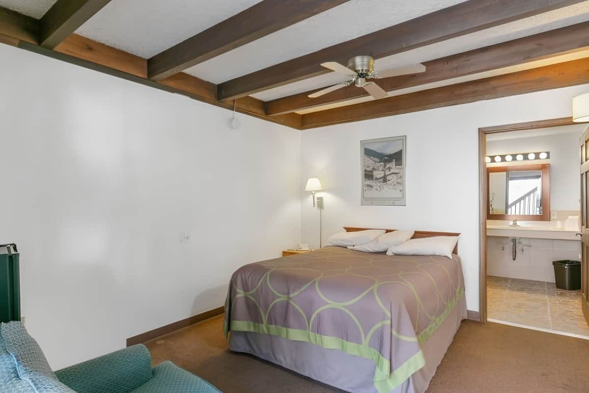 A bedroom with a bed, a ceiling fan, a nightstand, a wall-mounted lamp, and a bathroom with a mirror and sink.