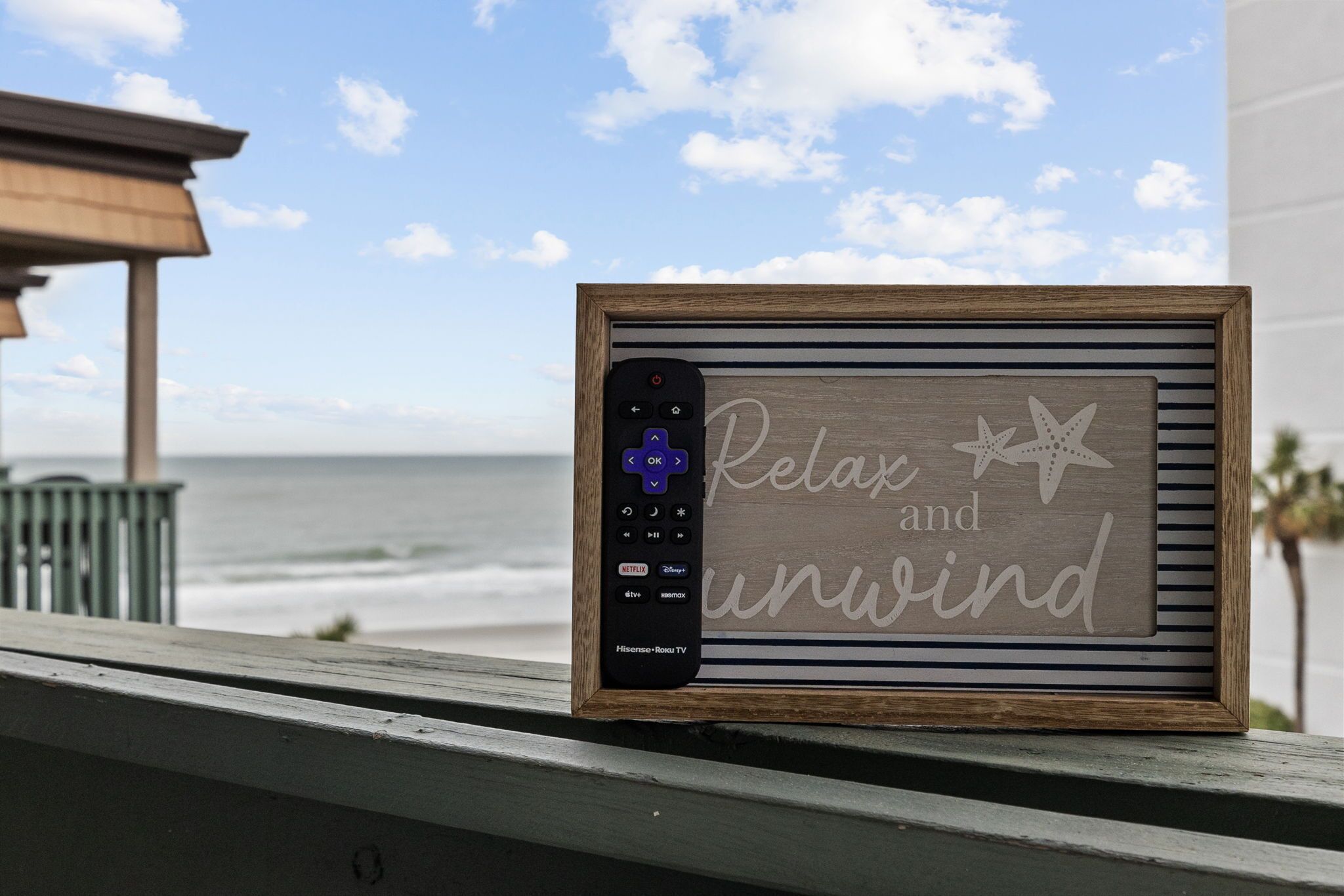 A framed sign with a remote control and the words "Relax and unwind" on a balcony overlooking the ocean.
