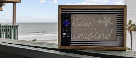 A framed sign with a remote control and the words "Relax and unwind" on a balcony overlooking the ocean.