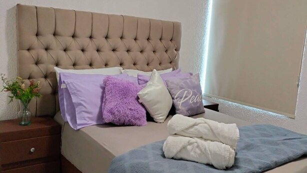 A bedroom with a tufted headboard, a bed with a blue blanket, and a nightstand with a vase of flowers. - Autódromo Apartment, Foro Solo, Cdmx Airport (Ciudad de México)