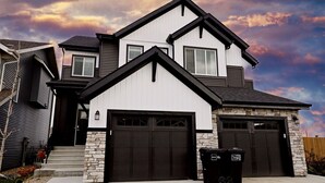 Une maison à deux étages avec une façade en pierre, un garage à deux portes et un petit jardin devant la maison, où se trouve une poubelle.
