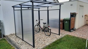 Ein überdachter Außenbereich mit einem Fahrrad, einer grünen Mülltonne und einer hölzernen Bank.