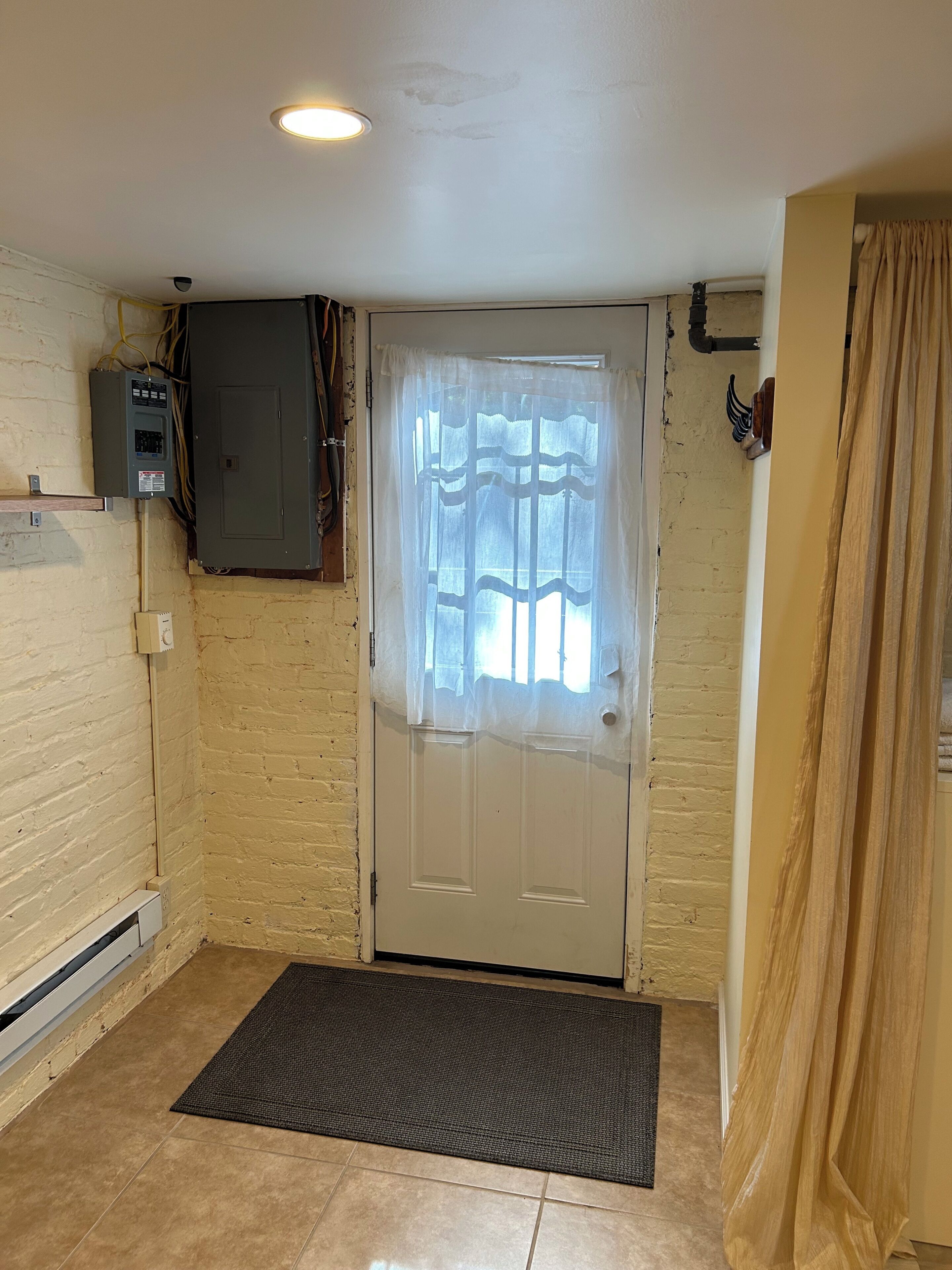 Une pièce avec une porte, un coffret électrique, une fenêtre avec des rideaux, un tapis et un luminaire fixé au mur.