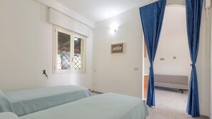 Une chambre avec deux lits, un banc, une fenêtre ornée d’une grille en métal et des rideaux bleus.