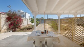 Un espace de restauration en plein air avec une table dressée pour deux personnes, entourée de chaises blanches, sous une pergola blanche.