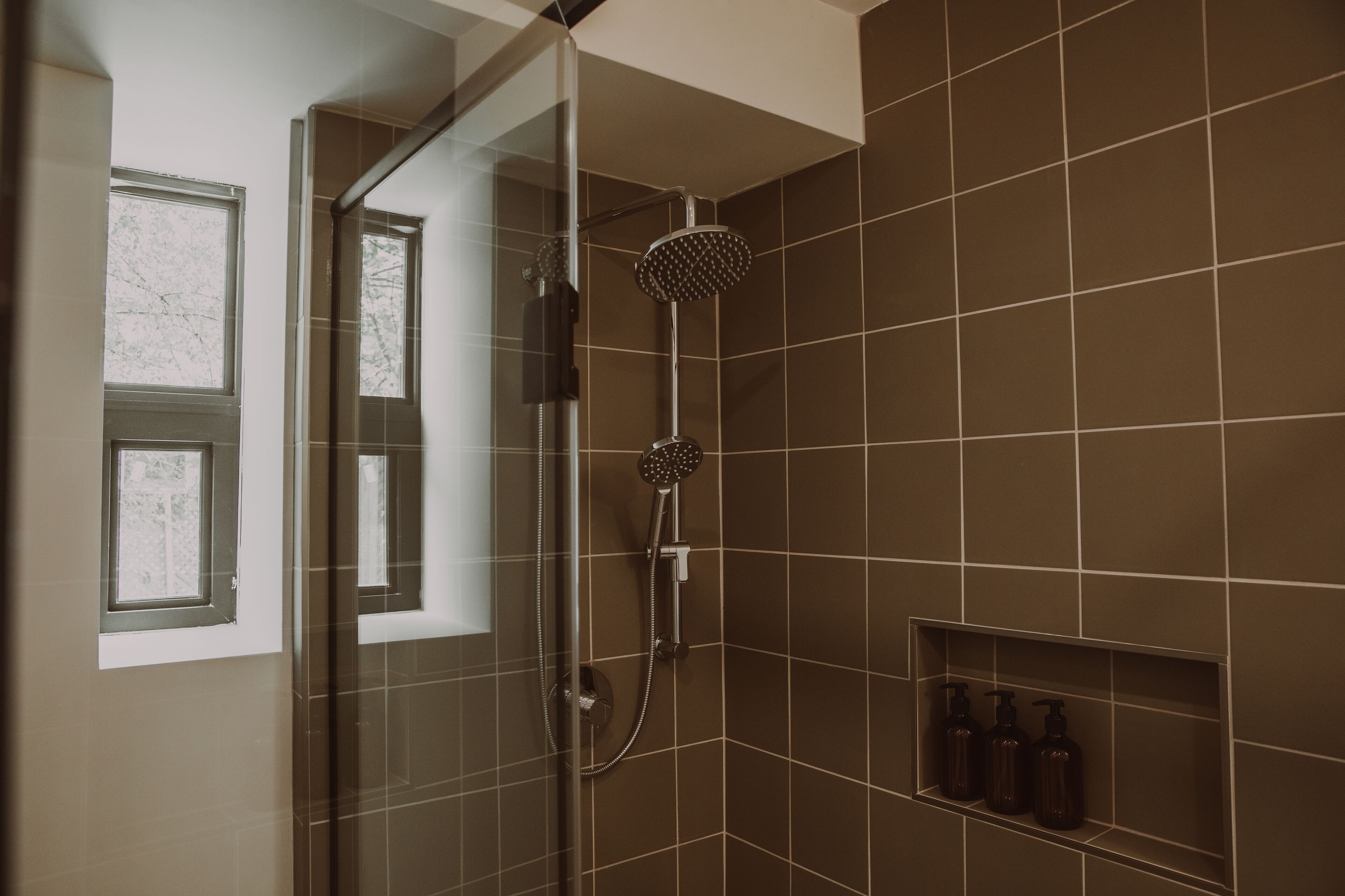 A shower with a handheld showerhead and a wall-mounted control. There is a window with a view of trees and a shelf with toiletries.