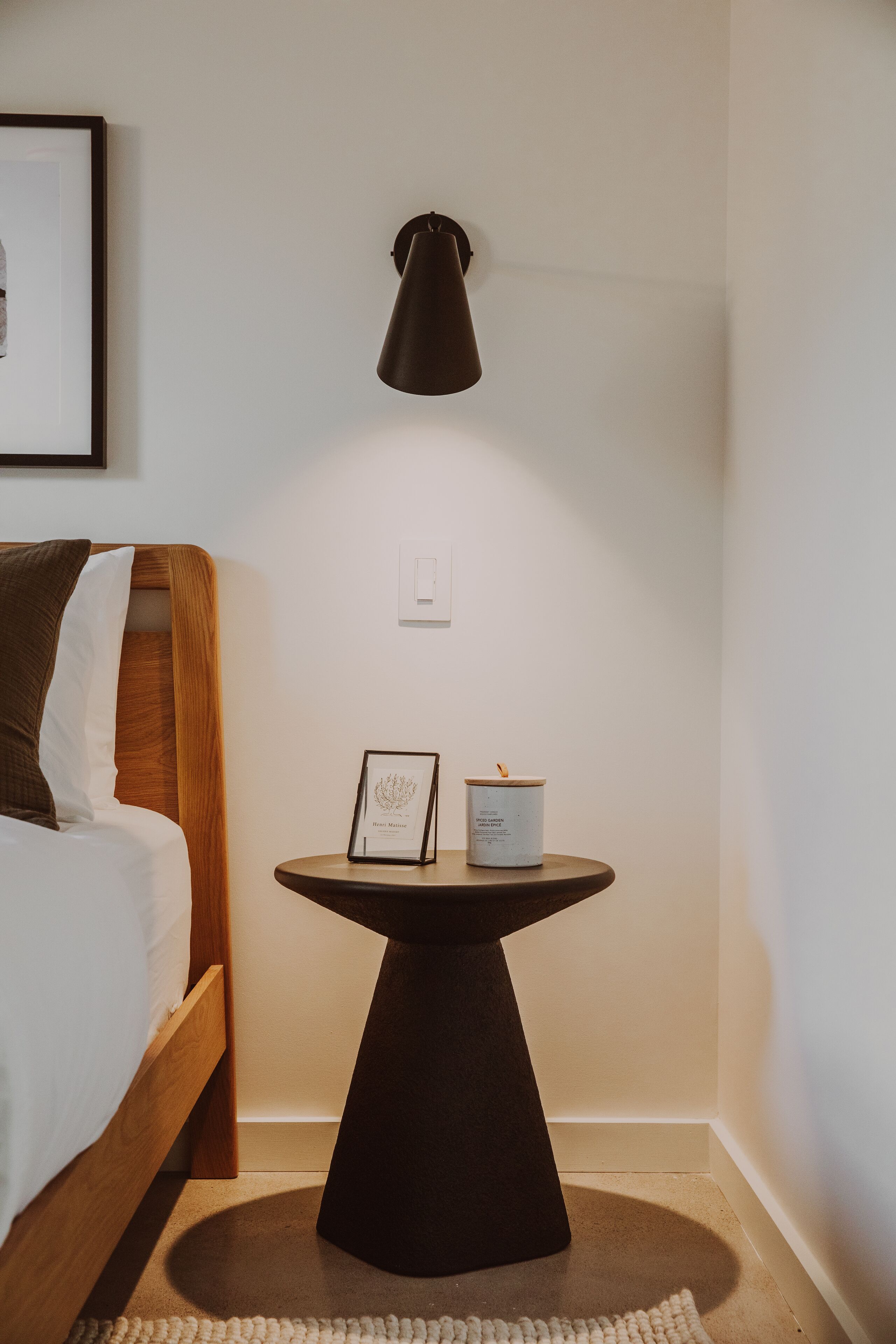 Un lit bien fait, avec une tête de lit en bois, une petite table ronde sur laquelle se trouvent une bougie et un objet encadré, et une lampe fixée au mur.