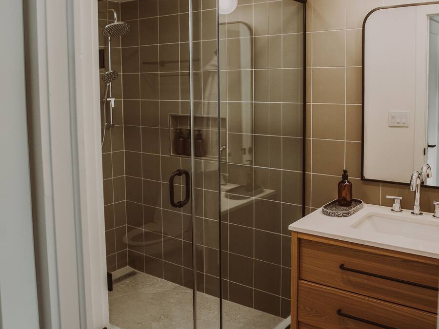 Un baño moderno con ducha de vidrio, tocador de madera con espejo y pared con azulejos color beige.