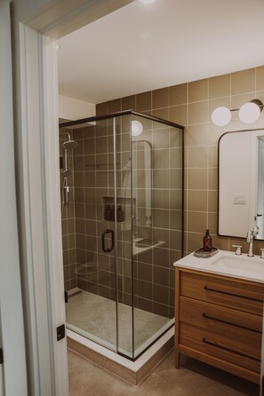 Un baño moderno con ducha de vidrio, tocador de madera con espejo y pared con azulejos color beige.