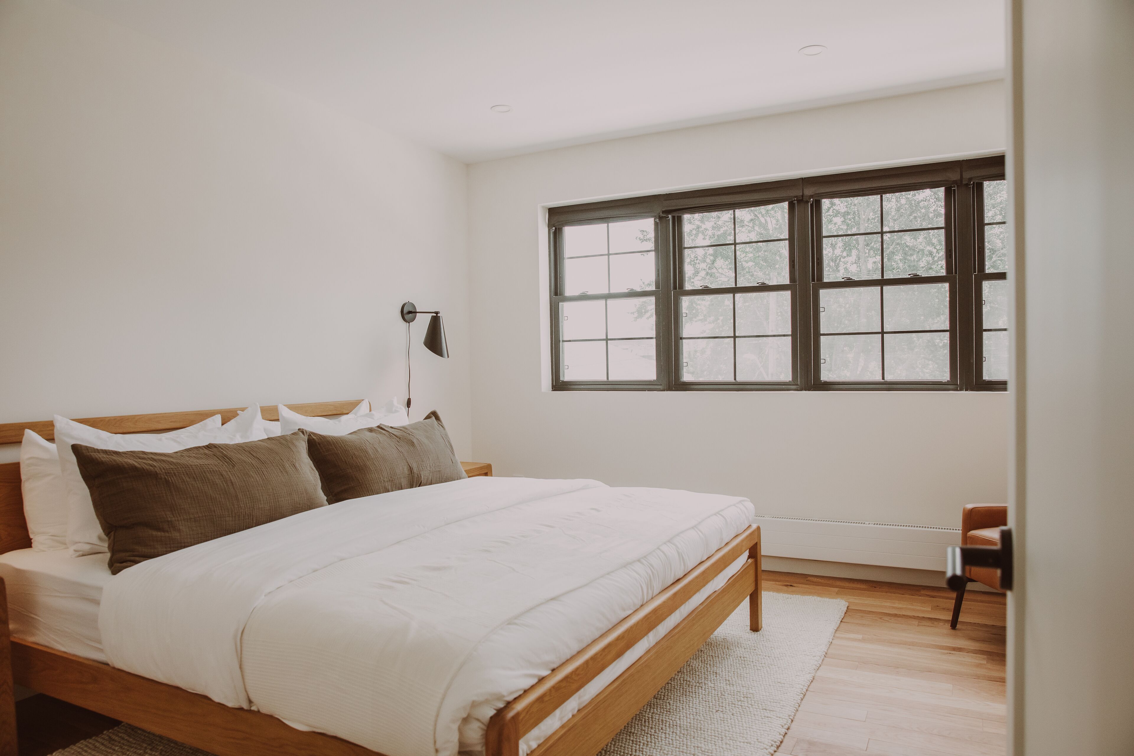 A bedroom with a wooden bed, white bedding, a large window, and a chair.