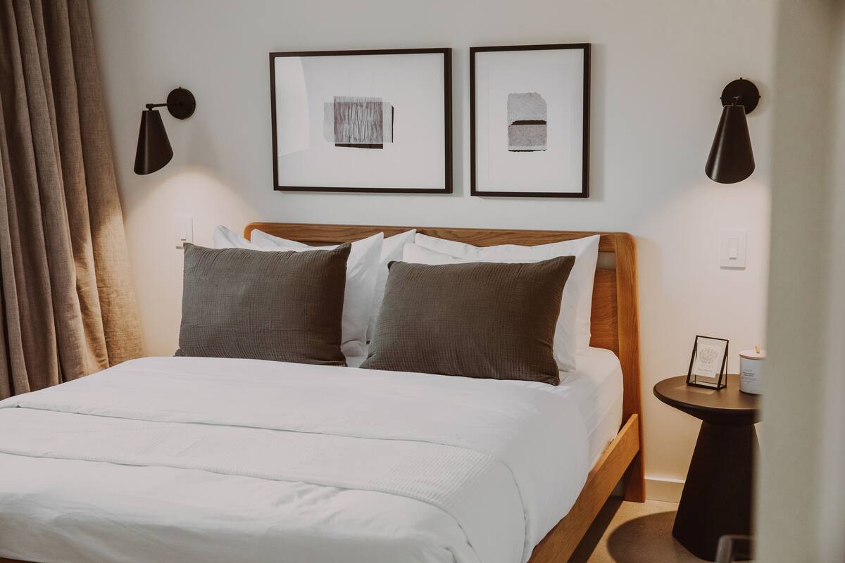 Un lit bien fait, avec du linge de lit blanc et gris, deux œuvres d’art encadrées au mur, et une petite table ronde sur laquelle se trouvent une bougie et une photographie encadrée.