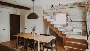 Un coin repas avec une table et des chaises en bois, un luminaire suspendu et un escalier adossé à un mur en pierre.