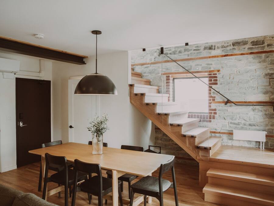 Un coin repas avec une table et des chaises en bois, un luminaire suspendu et un escalier adossé à un mur en pierre.