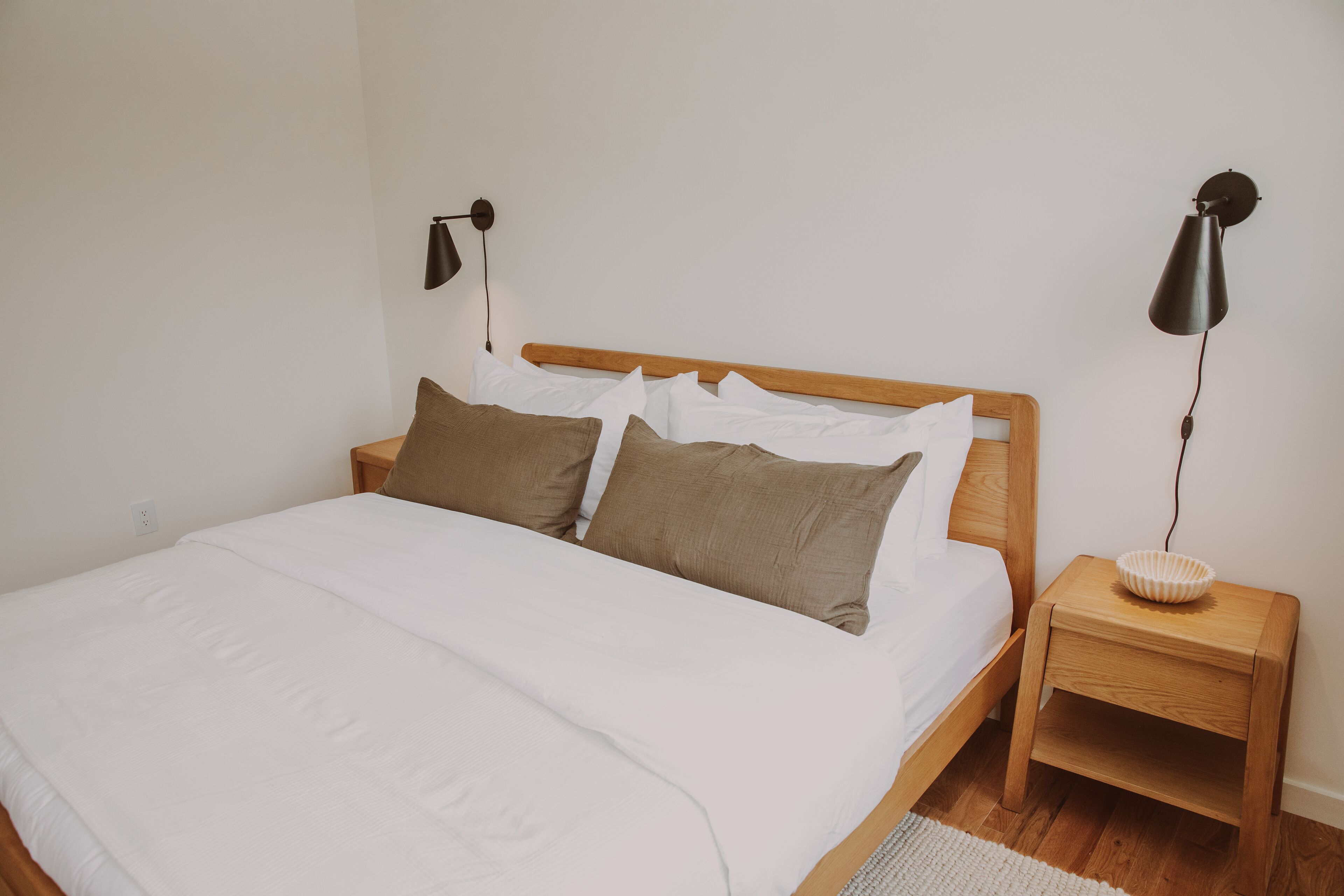 Una cama bien tendida con un cabecero de madera, dos mesitas de noche con lámparas y una alfombra blanca en el suelo.