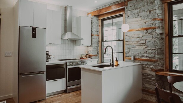 Une cuisine moderne avec des meubles blancs, un réfrigérateur en acier inoxydable, une cuisinière à gaz et un mur en pierre.