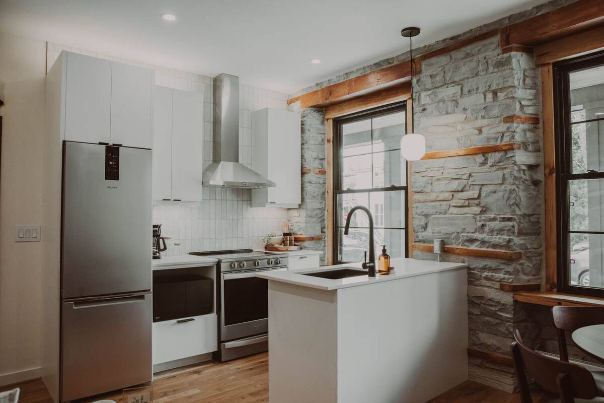 Une cuisine moderne avec des meubles blancs, un réfrigérateur en acier inoxydable, une cuisinière à gaz et un mur en pierre.
