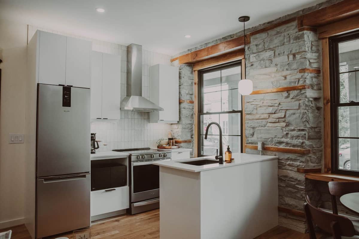Una cocina moderna con armarios blancos, un refrigerador de acero inoxidable, una estufa a gas y una pared de piedra.