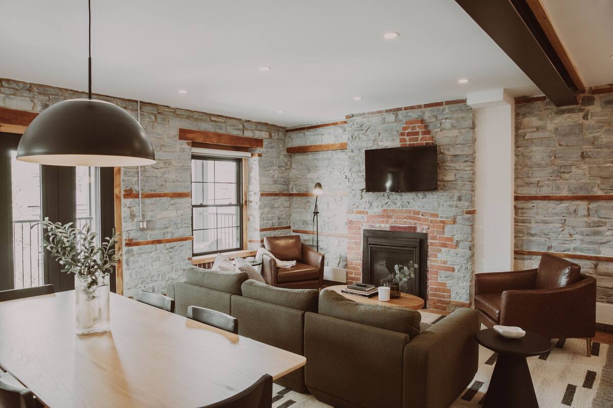 Un salon moderne avec un mur en pierre, une cheminée, des canapés en cuir et un coin repas doté d’une table en bois.