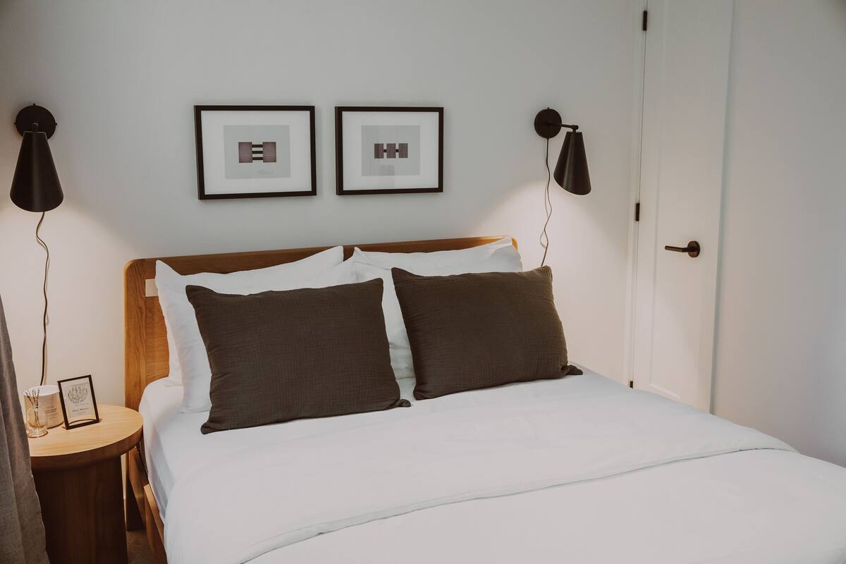 Un lit bien fait, avec une literie blanche et marron, deux tableaux encadrés au mur et une table de chevet avec une lampe.