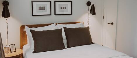 Una cama bien hecha con ropa de cama blanca y marrón, dos cuadros en la pared y una mesita de noche con una lámpara.