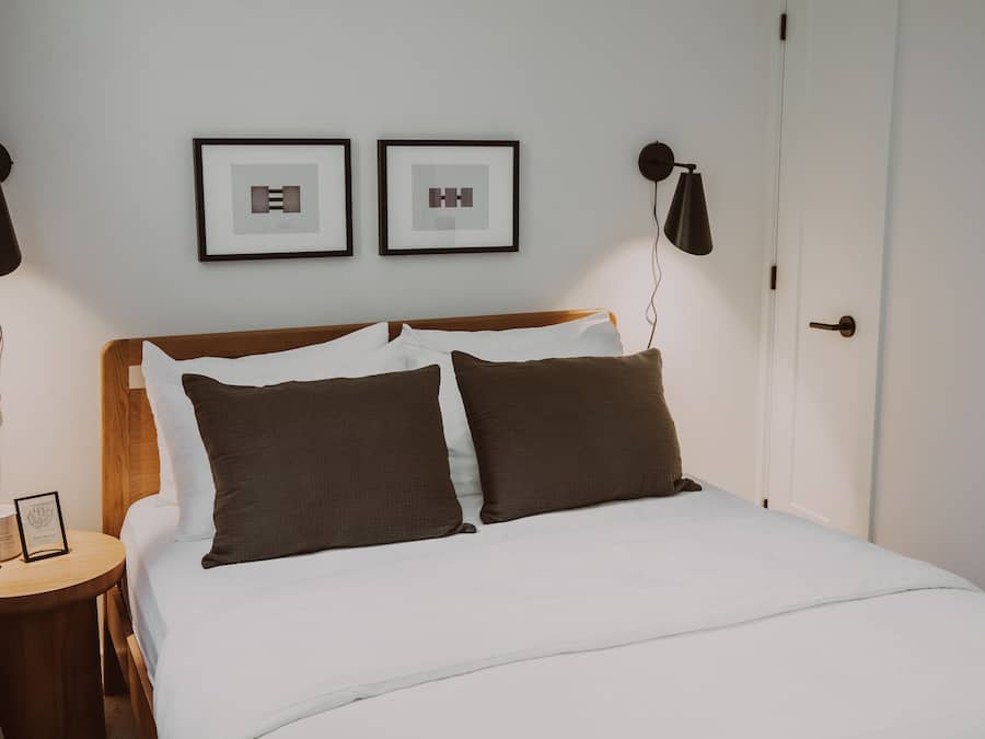 Un lit bien fait, avec une literie blanche et marron, deux tableaux encadrés au mur et une table de chevet avec une lampe.