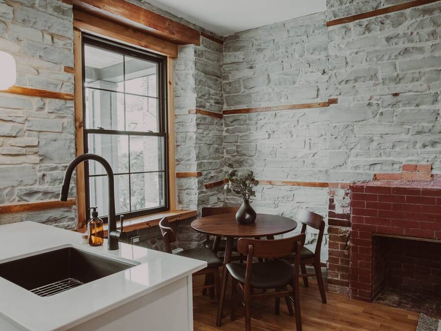 A kitchen with a stone wall, a window, a sink, and a dining area with chairs and a table.