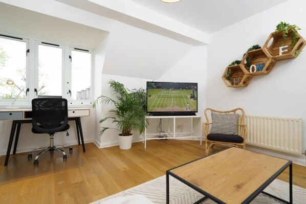 Un salon moderne avec un plancher en bois, un fauteuil de bureau noir, une table basse en bois, un téléviseur posé sur un meuble blanc, une plante en pot et un fauteuil en osier avec un coussin.