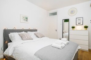 Un lit bien fait, avec une literie blanche et grise, une tête de lit grise et une table de chevet avec une lampe.