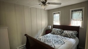 A bedroom with a wooden bed, a ceiling fan, two windows with patterned curtains, and a white wardrobe. - Quiet Cottage in the heart of Burr Oak State Park. (Glouster)