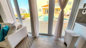 A room with a view of a swimming pool, a white sofa with teal pillows, and a trash bin. - Tropical Retreat Stay-Spacious 5BR Oasis with Pool, Solar Backup, for 20 Guests! (caimital alto)