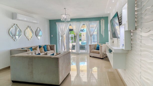 A spacious living room with a large sofa, a television, and a view of the outdoor area. - Tropical Retreat Stay-Spacious 5BR Oasis with Pool, Solar Backup, for 20 Guests! (caimital alto)