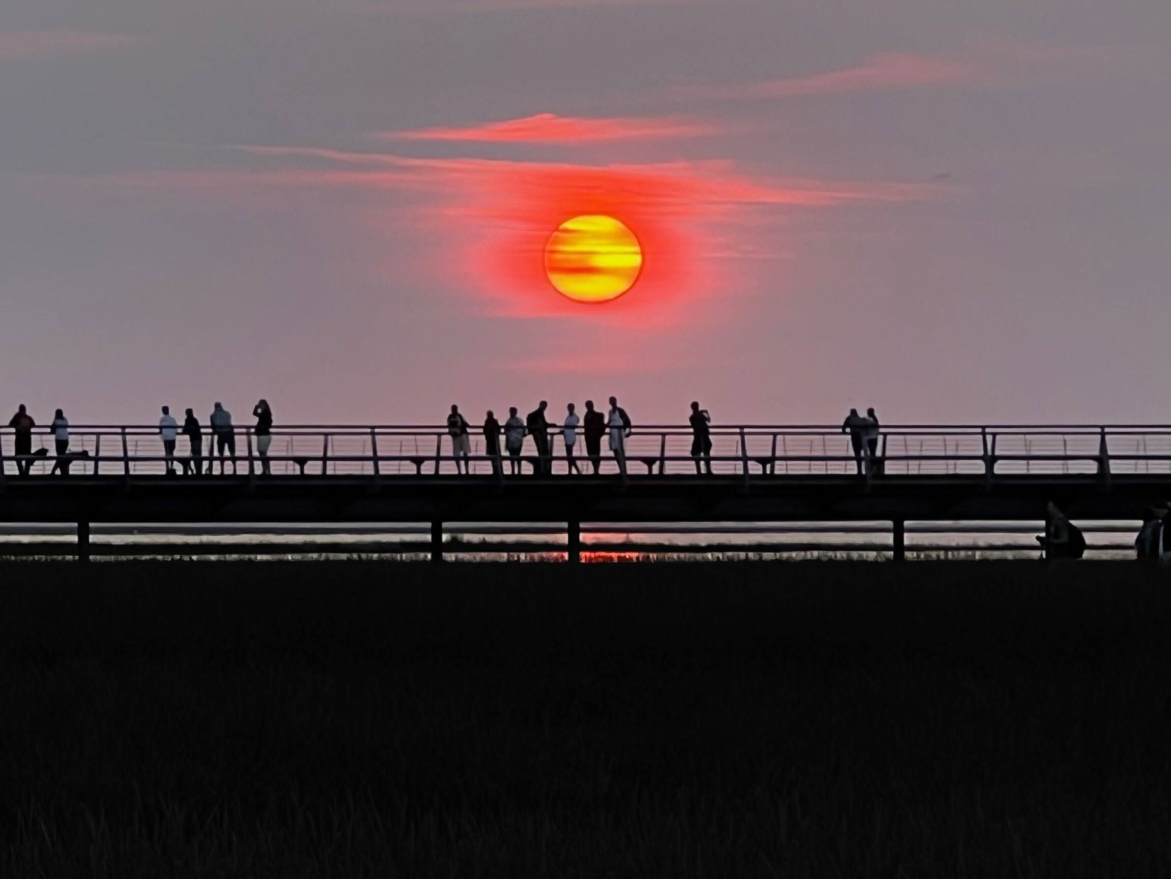 Un atardecer sobre un cuerpo de agua, con siluetas de personas en un puente contra el cielo.