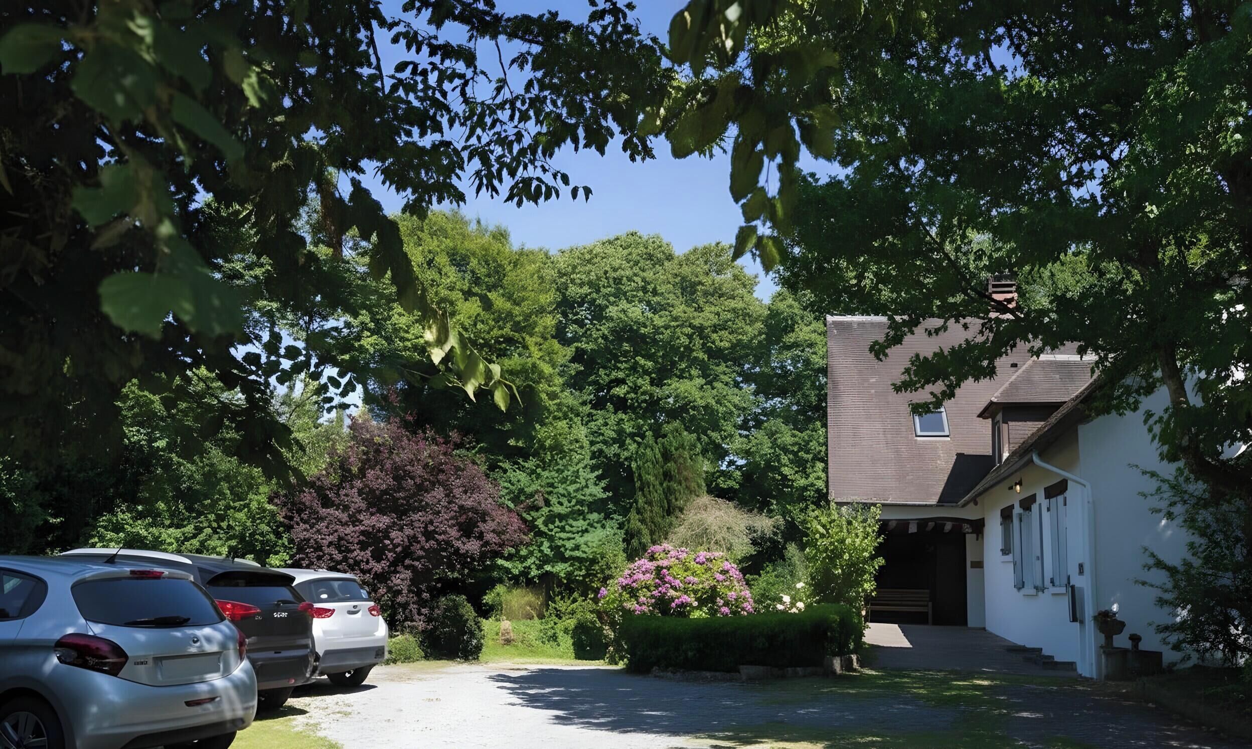 A residential street with parked cars, a house with a sloped roof, and lush greenery.