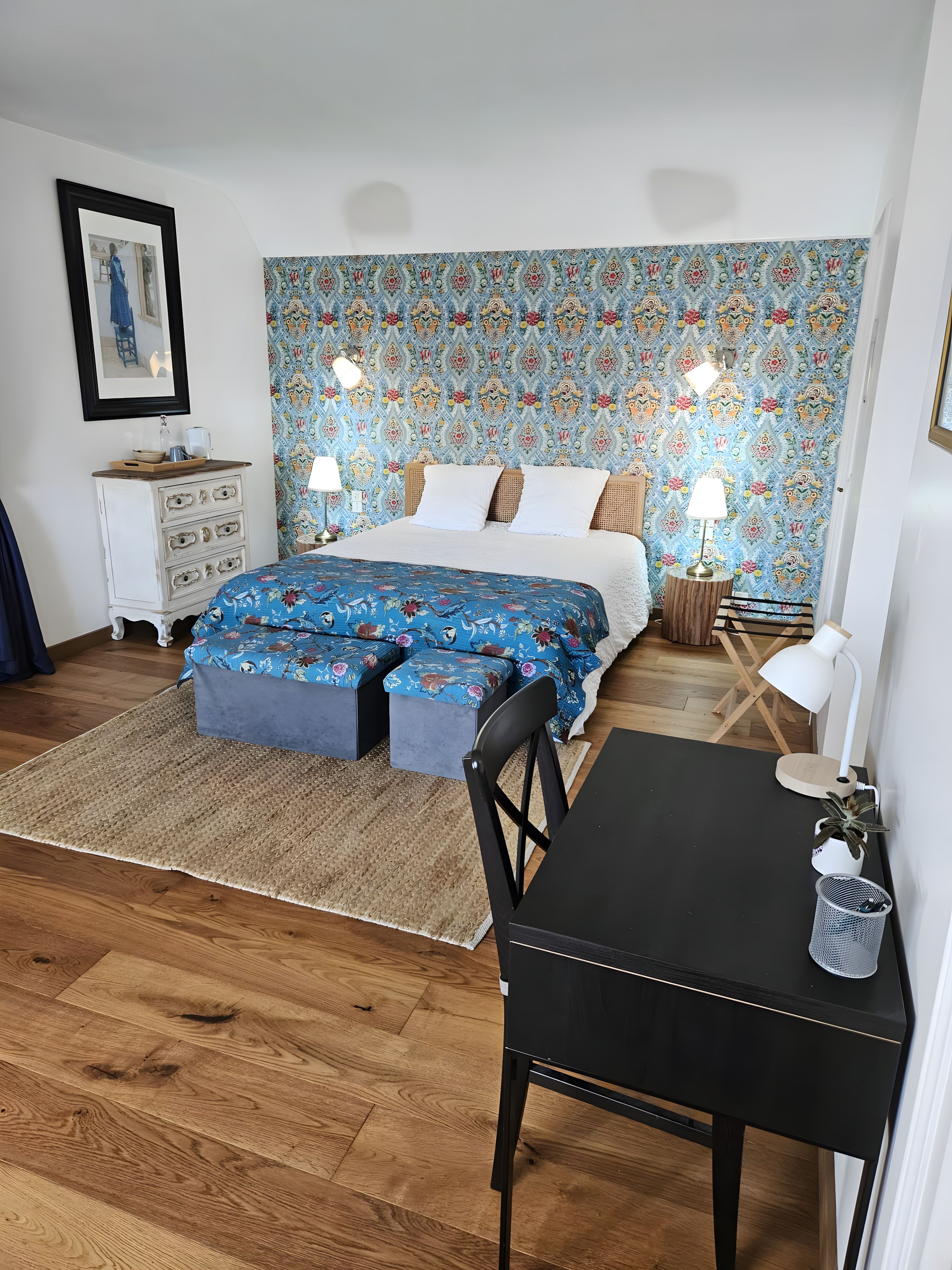 A bedroom with a bed, bedside tables, a dresser, a chair, a table, and a patterned wall.
