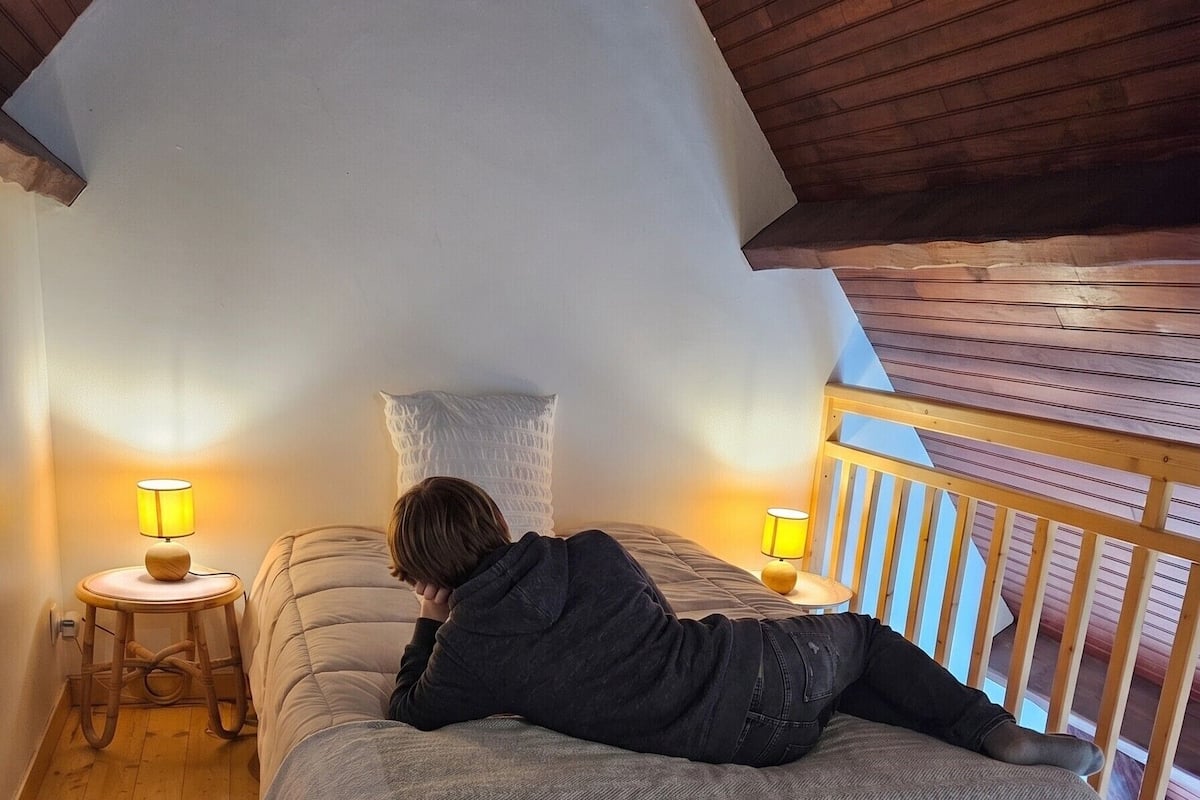 A bedroom with a sloped ceiling, a bed with a grey blanket, two bedside lamps, and a wooden railing.