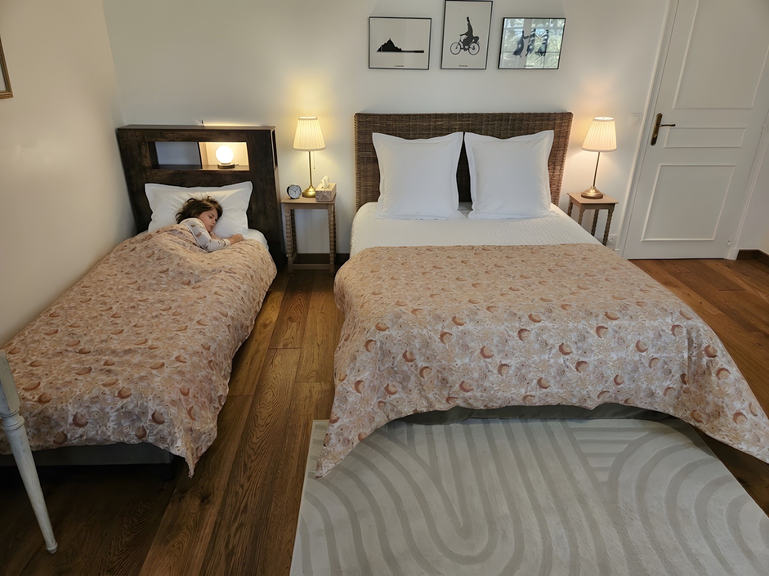 A hotel room with two beds, a wooden floor, a small bedside table with a lamp, and framed pictures on the wall.