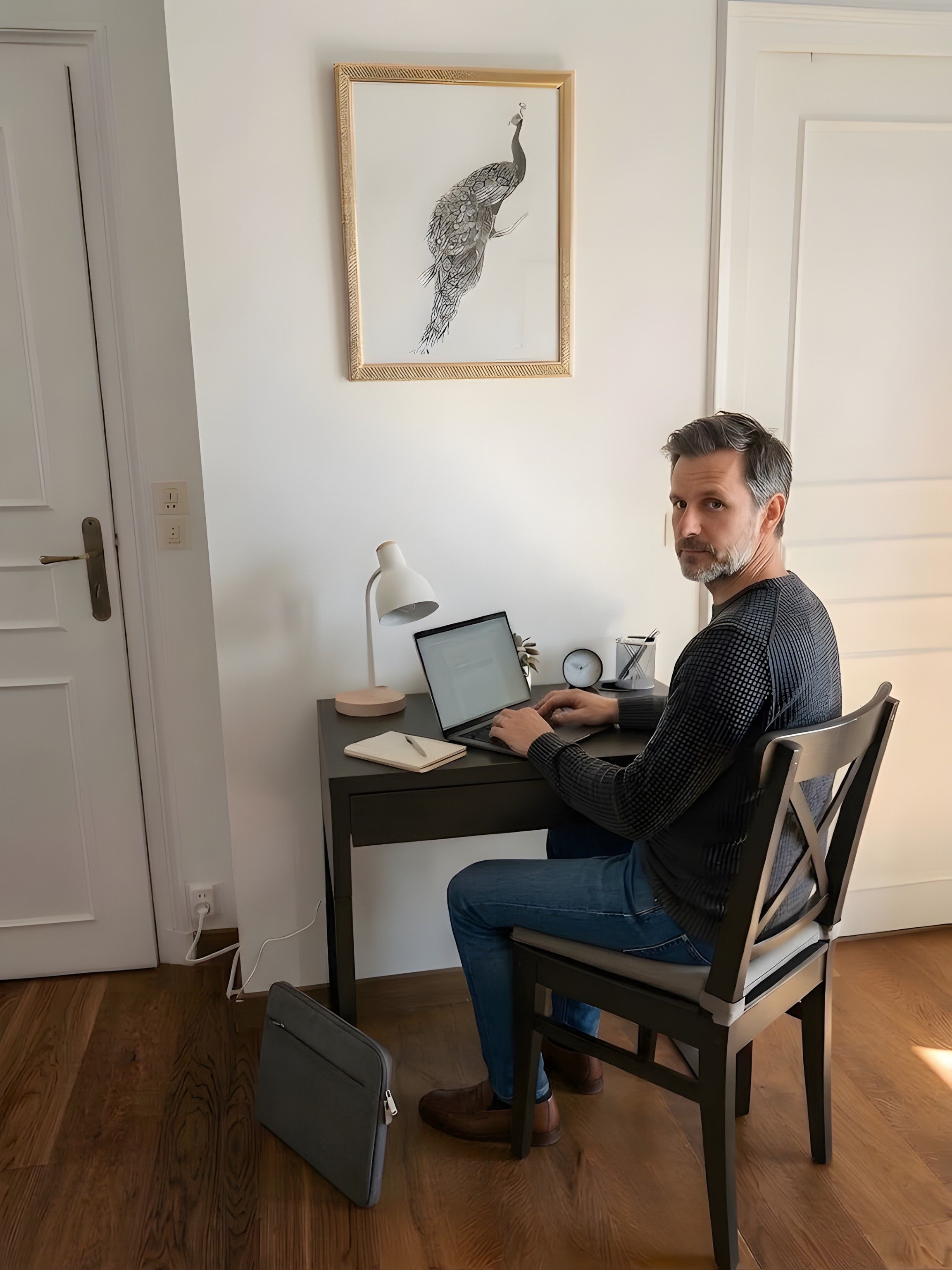 Eine Person sitzt an einem Schreibtisch mit einem Laptop, einem Notizbuch und einem Stift in einem Raum mit Holzböden und einem gerahmten Bild an der Wand.
