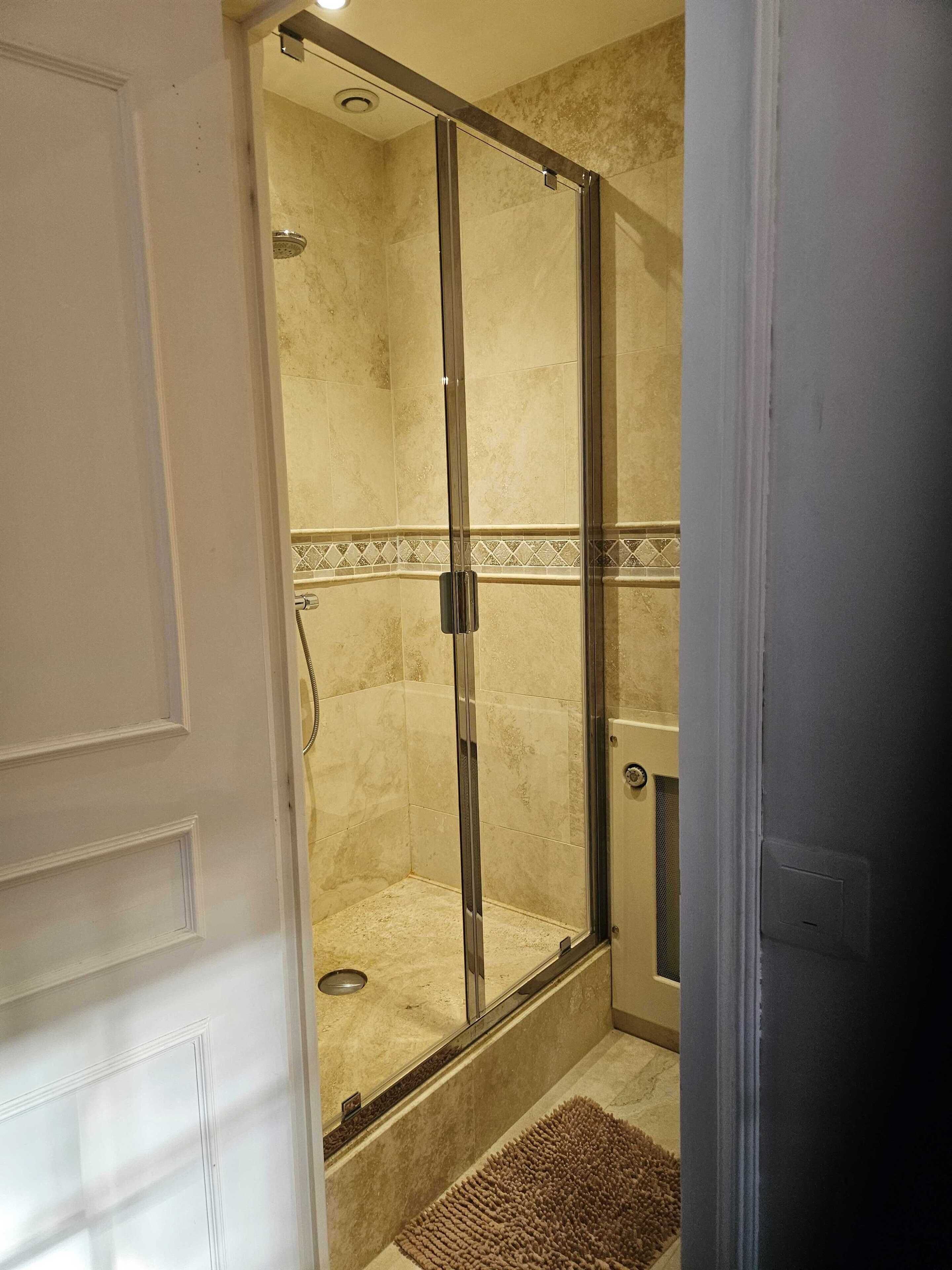 Une douche à l’italienne avec une porte en verre, des murs carrelés de couleur beige et un petit tapis sur le sol.