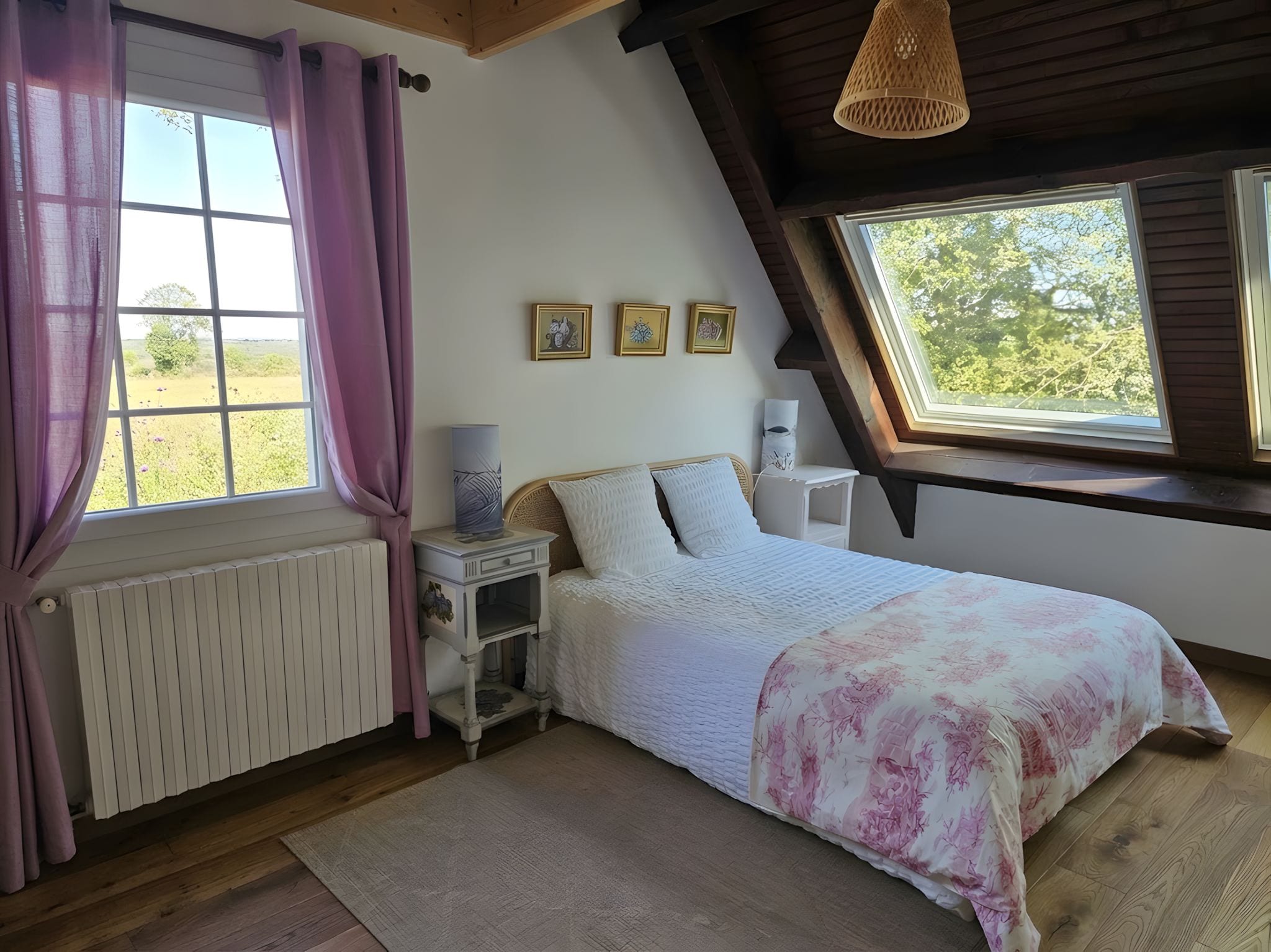 Un dormitorio con techo inclinado, una cama con colcha estampada, una mesita de noche con lámpara, un radiador y una ventana con cortinas.