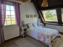 Un dormitorio con techo inclinado, una cama con colcha estampada, una mesita de noche con lámpara, un radiador y una ventana con cortinas.