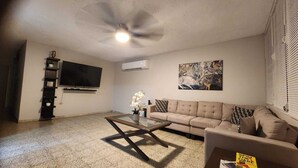 A living room with a beige sofa, a glass coffee table, a TV mounted on the wall, and an air conditioner. - Puerto Rico: 5-Minute Airport / Parking / Carolina, San Juan. (Carolina)